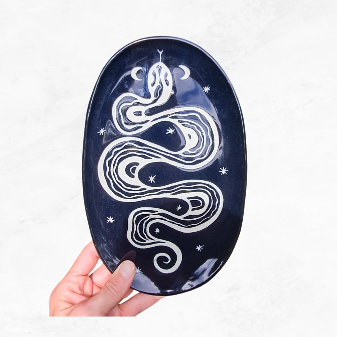 Ceramic oval plate, decorated with one large flowing snake, crescent moons and stars, black and white in colour.