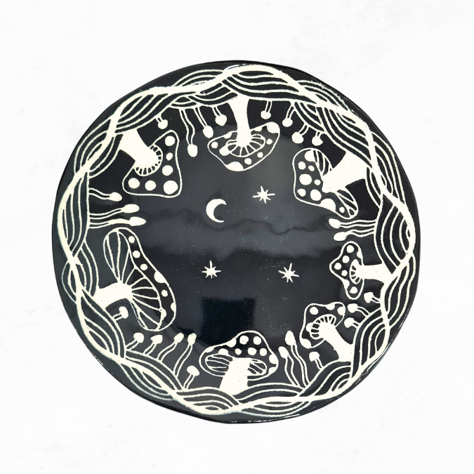 Ceramic plate decorated with crescent moon, stars in the middle and mushrooms around the edge, black and white in colour.