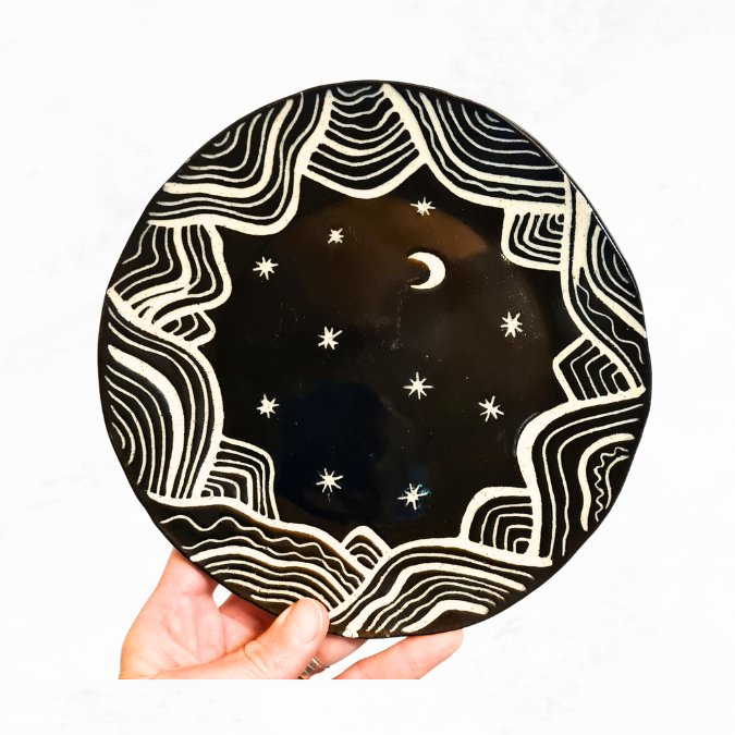 Ceramic plate decorated with crescent moon, stars and lines around the edge that represent mountains, black and white in colour.